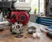 Carburator defect Honda GX analizat pe bancul de lucru, lângă un motor demontat